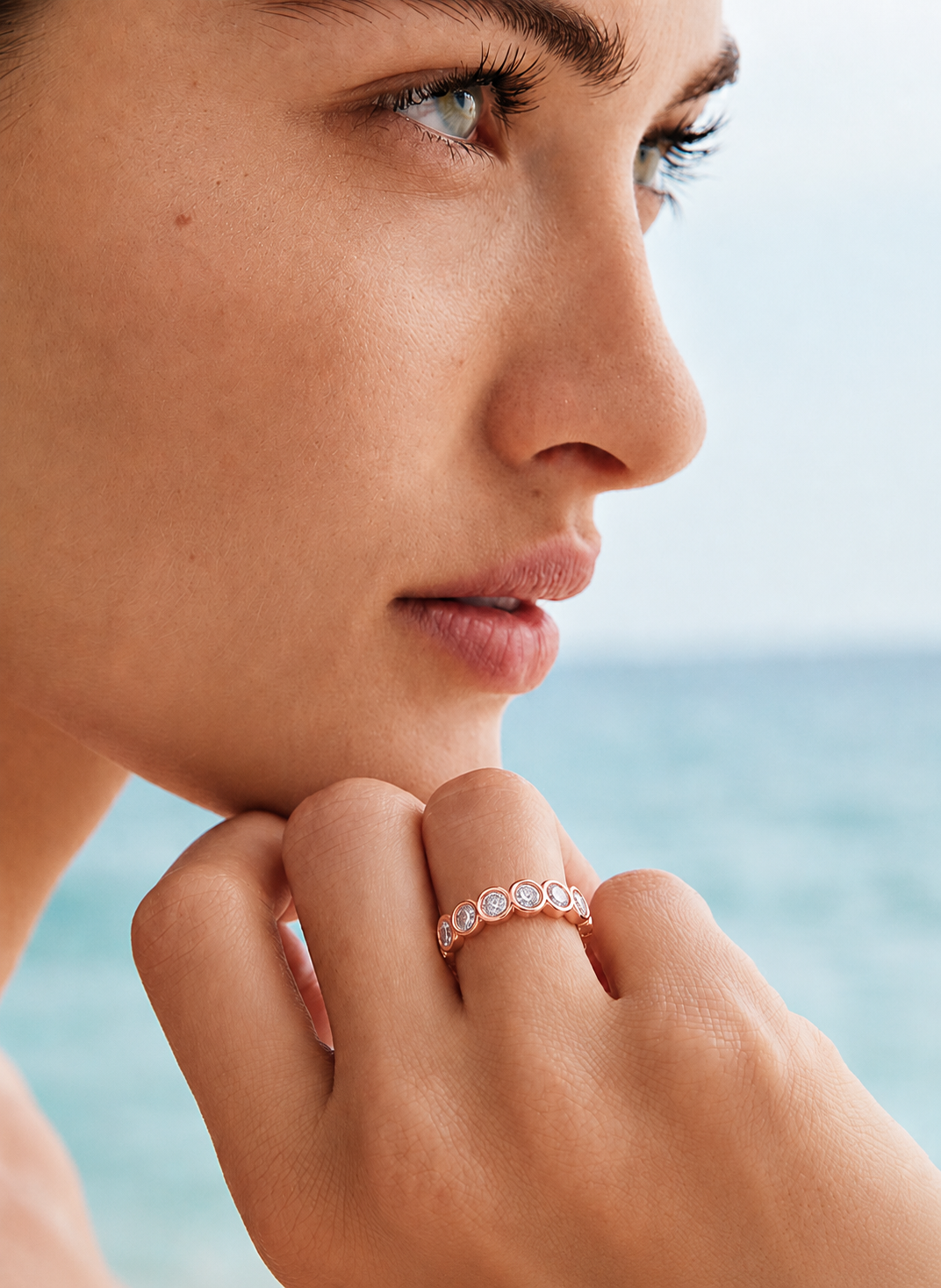 color_10K_RG, color_14K_RG &&	A close-up lifestyle shot of a woman's face and hand resting near her chin, wearing a delicate rose gold band ring with small clear round-cut gemstones, set against a soft-focus background of blue water and sky.