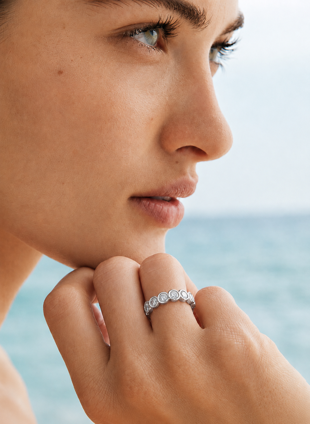 color_925_SIL,color_935_ARG,color_10K_WG, color_14K_WG &&	
A close-up lifestyle shot of a woman's face and hand, wearing a delicate silver ring with a row of small clear gemstones, set against a soft-focus background of the ocean and sky.