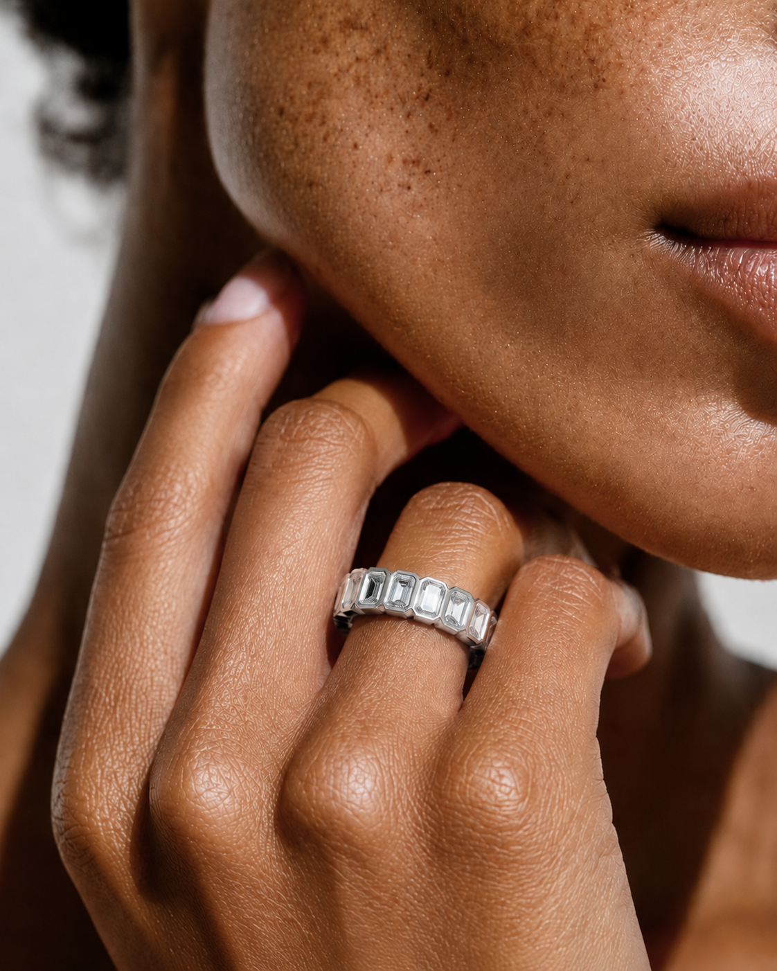 color_925_SIL,color_935_ARG,color_10K_WG, color_14K_WG &&	
A close-up of a person's hand wearing a silver band ring with a row of rectangular clear stones, set against a close-up of their face and neck.