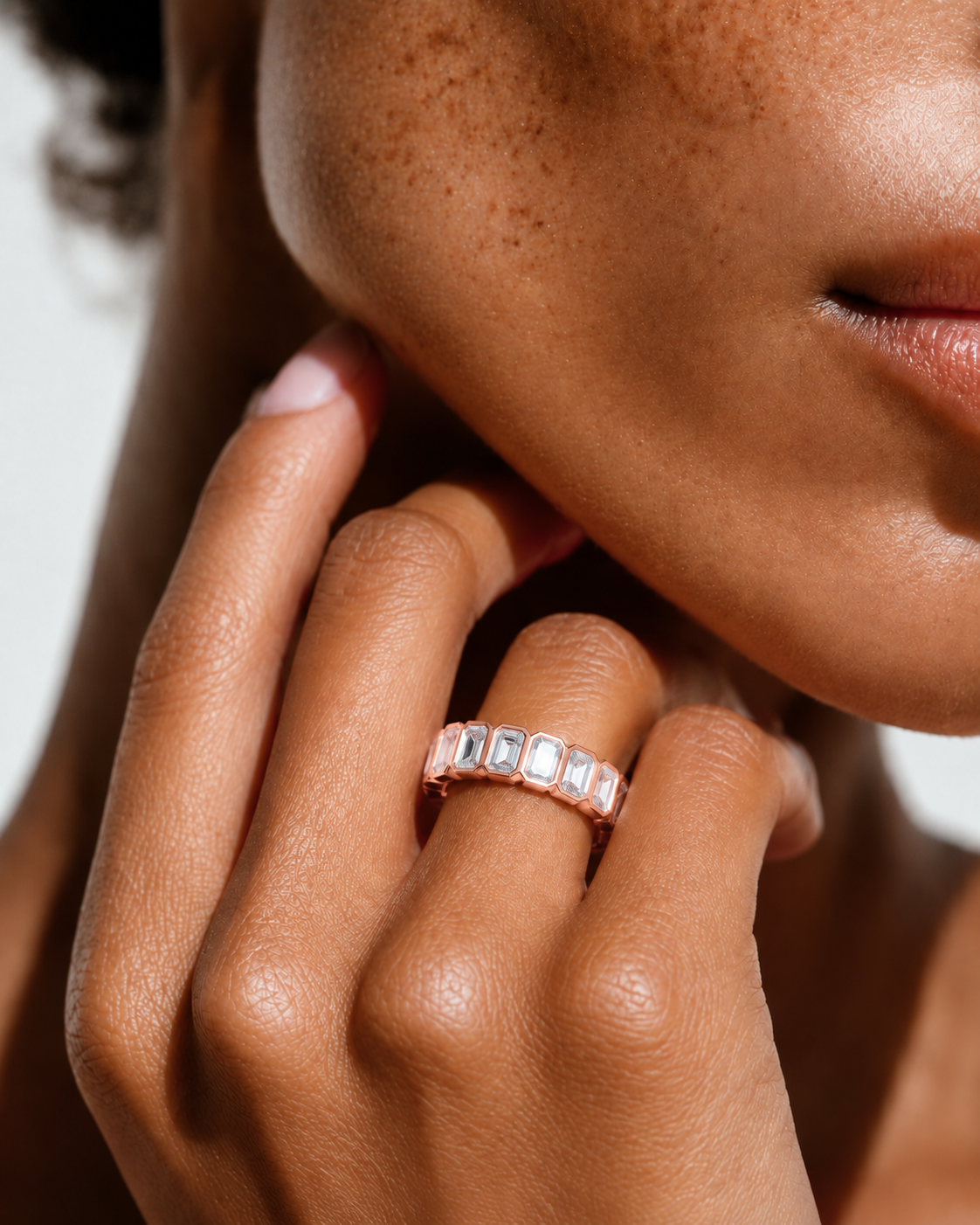 color_10K_RG, color_14K_RG &&	A close-up of a person's hand and lower face wearing a rose gold eternity ring with rectangular clear gemstones, set against a clean white background.