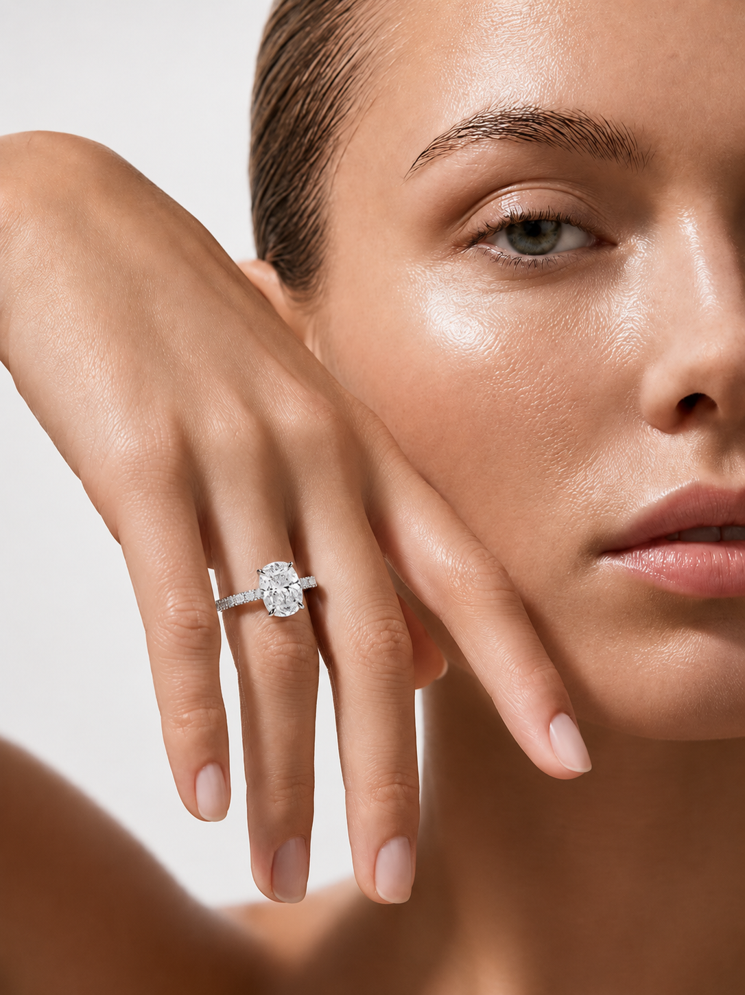 color_925_SIL,color_935_ARG,color_10K_WG, color_14K_WG &&A close-up lifestyle shot of a model wearing a diamond engagement ring on her finger, with her face and hand prominently framed against a soft light gray background.