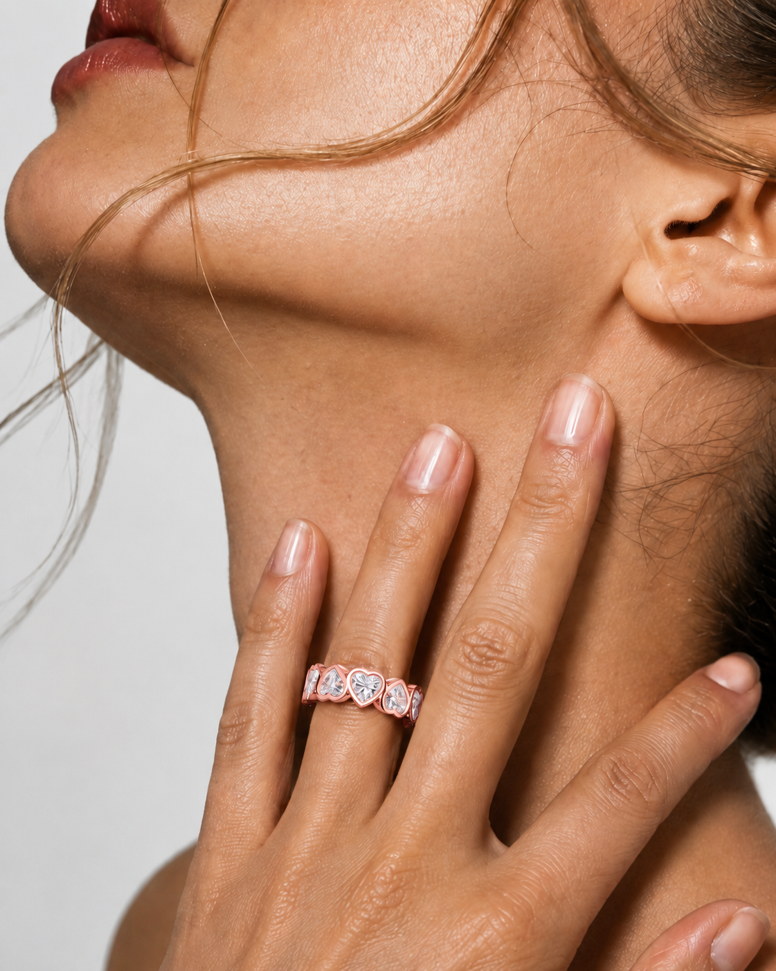 color_10K_RG, color_14K_RG &&	A close-up lifestyle shot of a model's hand resting on her neck, wearing a delicate rose gold band ring set with small clear heart-shaped gemstones.