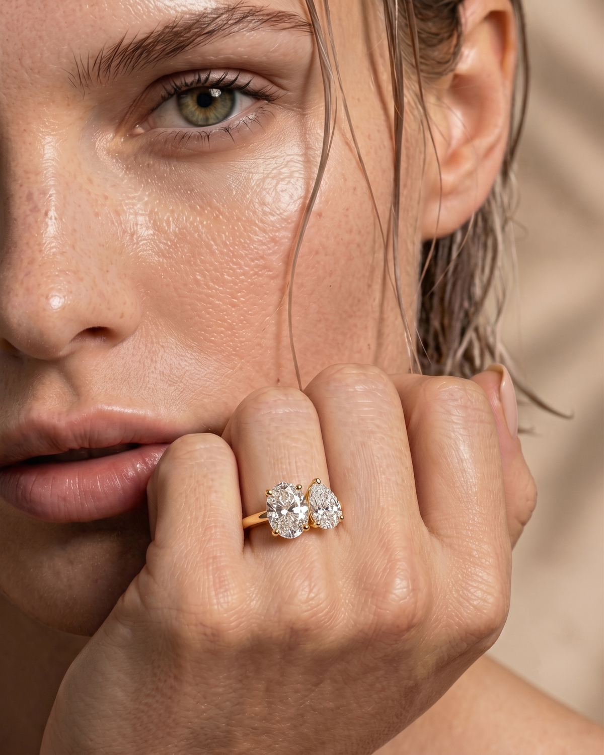color_10K_YG, color_14K_YG &&A close-up lifestyle shot of a model's face and hand, showcasing a gold ring with two round-cut clear gemstones in a heart-like arrangement.
