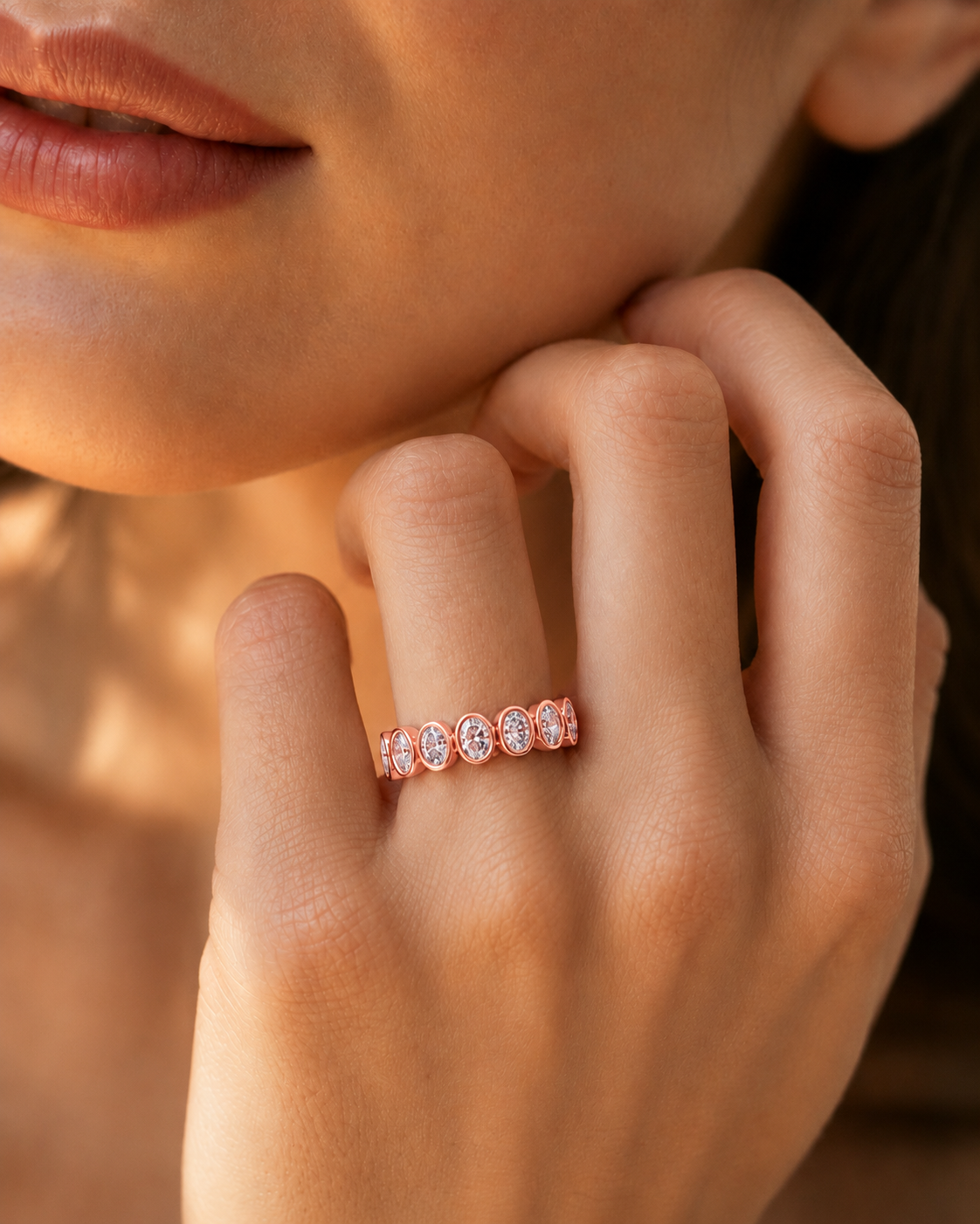 color_10K_RG, color_14K_RG &&	A close-up lifestyle shot of a woman's hand wearing a rose gold eternity ring with round-cut clear gemstones. The warm, golden-hour lighting highlights the delicate metal and sparkling stones, with the model's face and shoulder softly blurred in the background.