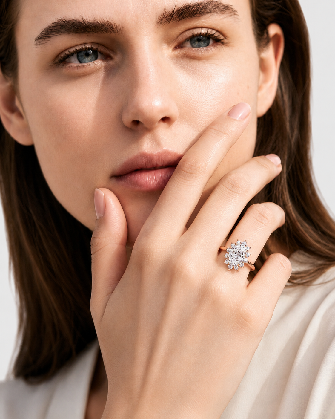 color_10K_RG, color_14K_RG &&A close-up lifestyle shot of a woman wearing a rose gold ring with a clear diamond cluster on her ring finger, set against a soft, light gray background.