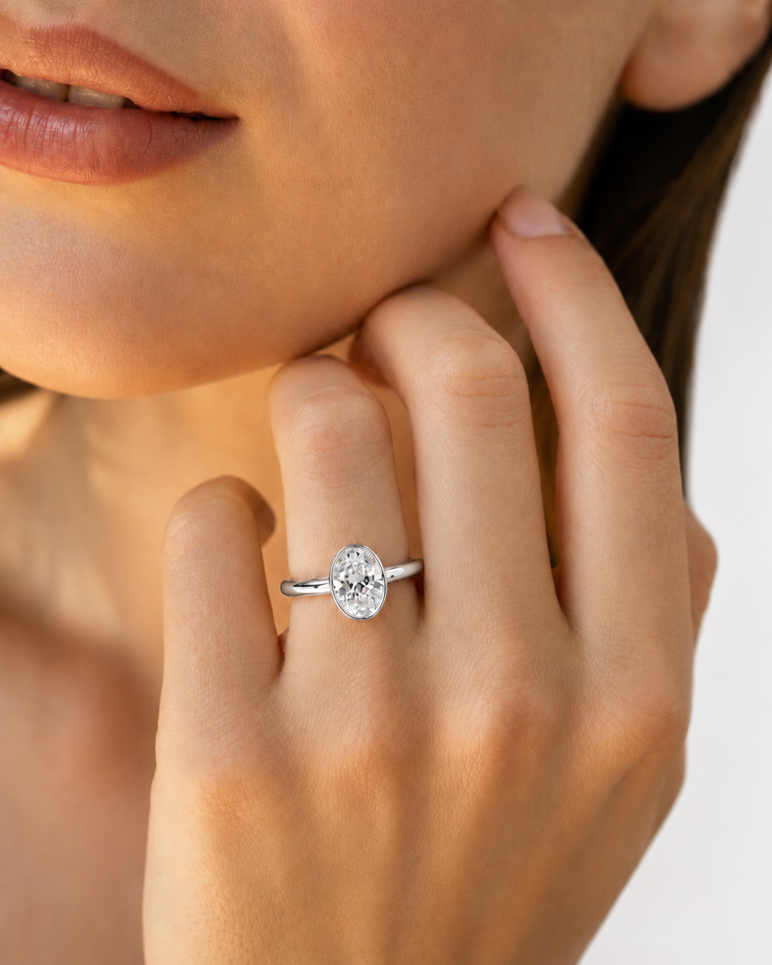 color_925_SIL,color_935_ARG,color_10K_WG, color_14K_WG &&A close-up lifestyle shot of a woman wearing a silver band ring with an oval-cut clear gemstone on her finger.