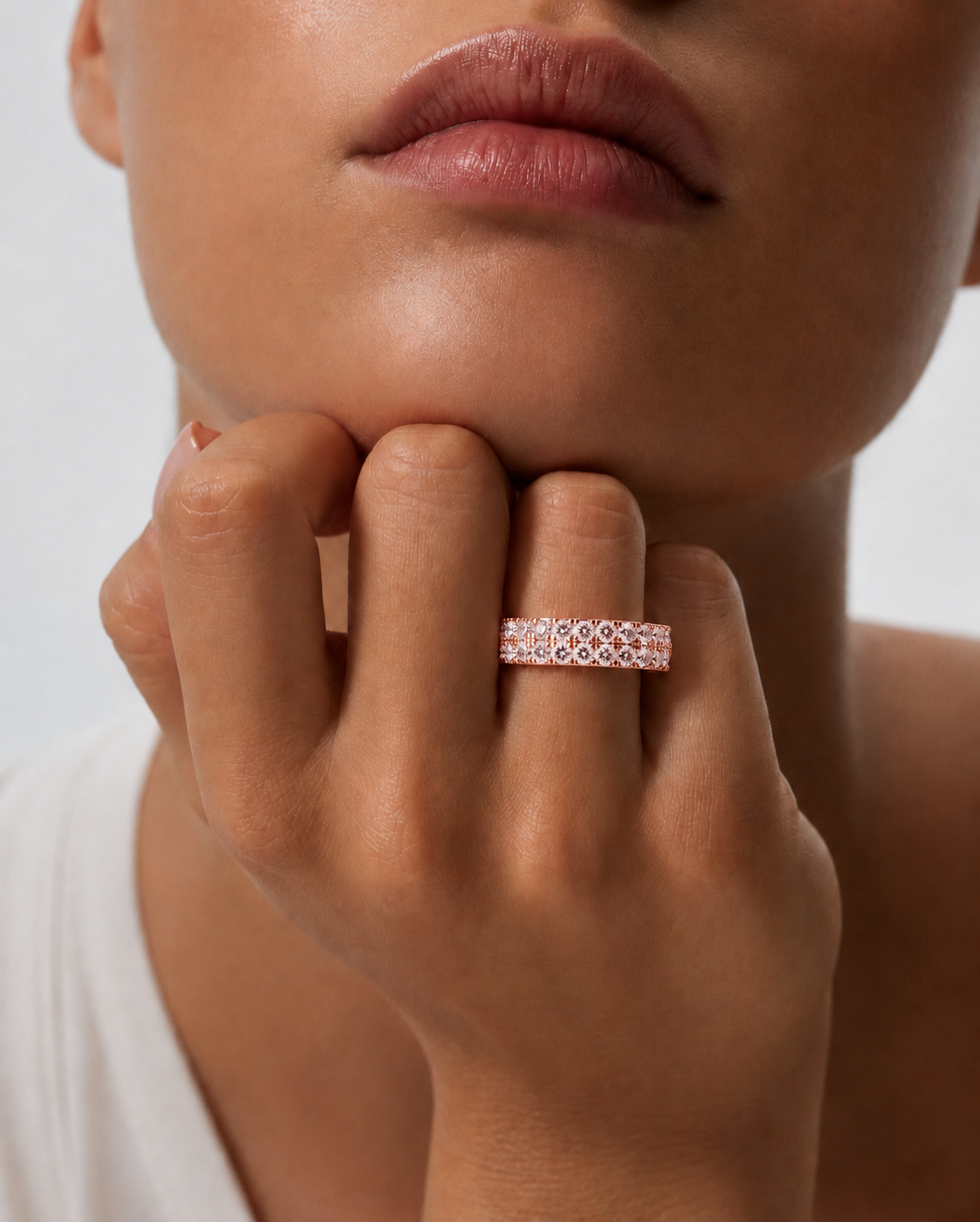 color_10K_RG, color_14K_RG &&	A close-up lifestyle shot of a model wearing a rose gold eternity ring with small clear gemstones on her finger, with her hand resting on her chin against a clean white background.