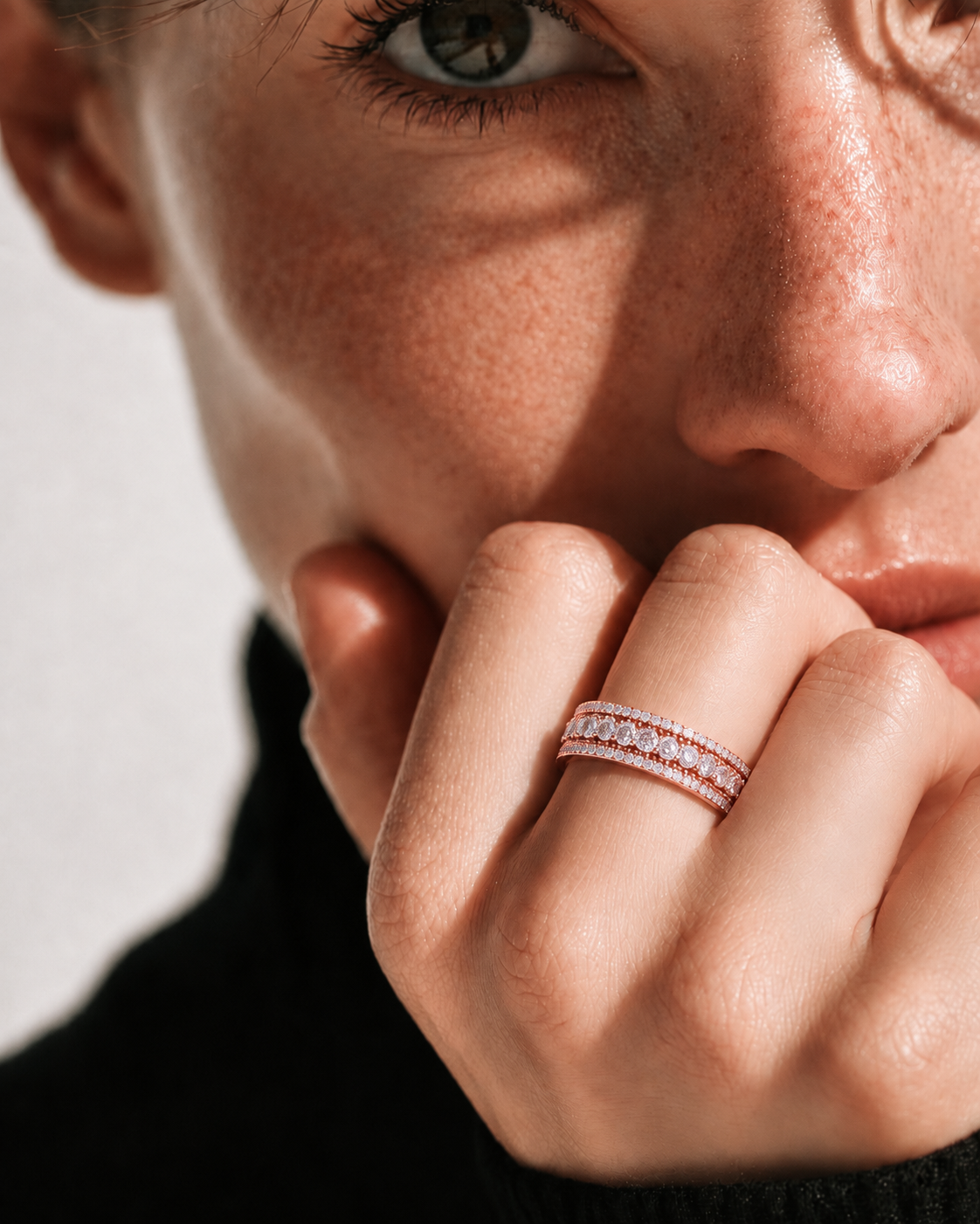 color_10K_RG, color_14K_RG &&	A close-up lifestyle shot of a model wearing a delicate rose gold band ring with pave-set clear stones. The ring is shown on the model's ring finger, with her hand resting near her chin against a neutral light background.