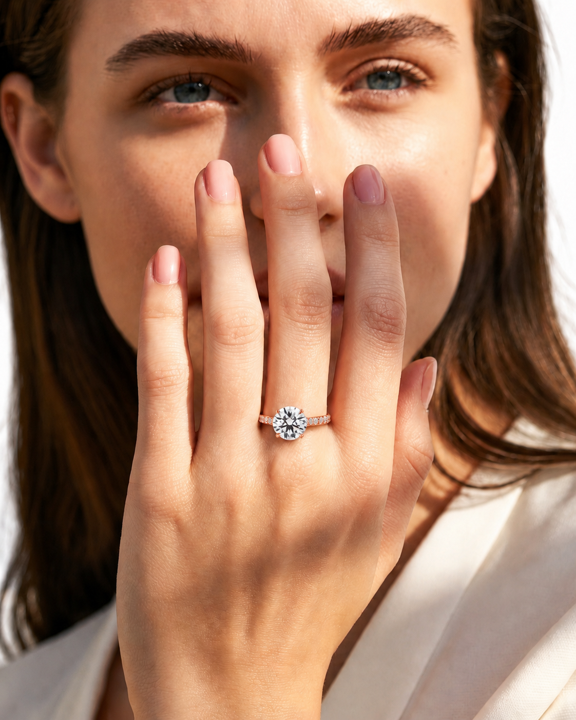 color_10K_RG, color_14K_RG &&A close-up lifestyle shot of a woman wearing a silver-toned diamond engagement ring on her finger, with her hand covering part of her face against a bright white background.