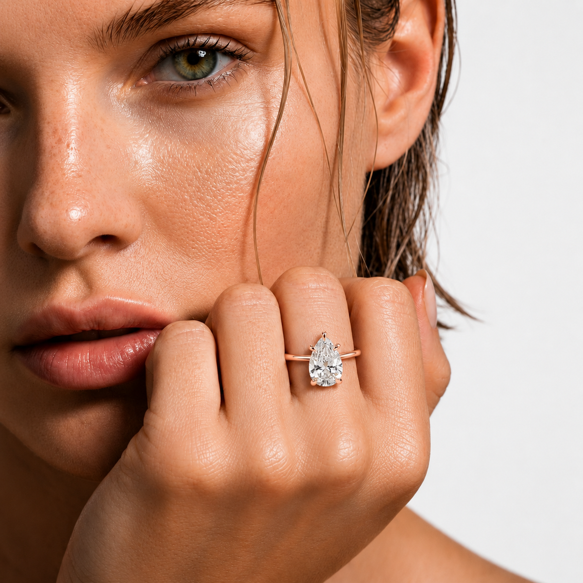 color_10K_RG, color_14K_RG &&A close-up lifestyle shot of a model wearing a pear-cut diamond ring on a slim rose gold band. The model's face and hand are shown in warm, direct lighting against a clean white background.