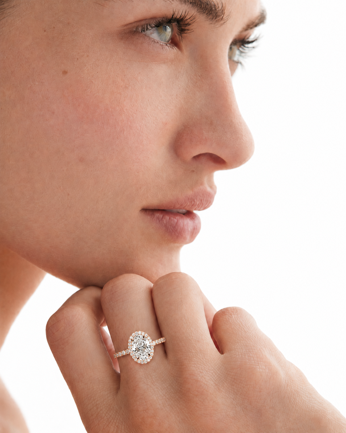 color_10K_RG, color_14K_RG &&A close-up side profile of a woman wearing a diamond engagement ring with a round-cut center stone and a halo setting on a slim band, shown against a bright white background.