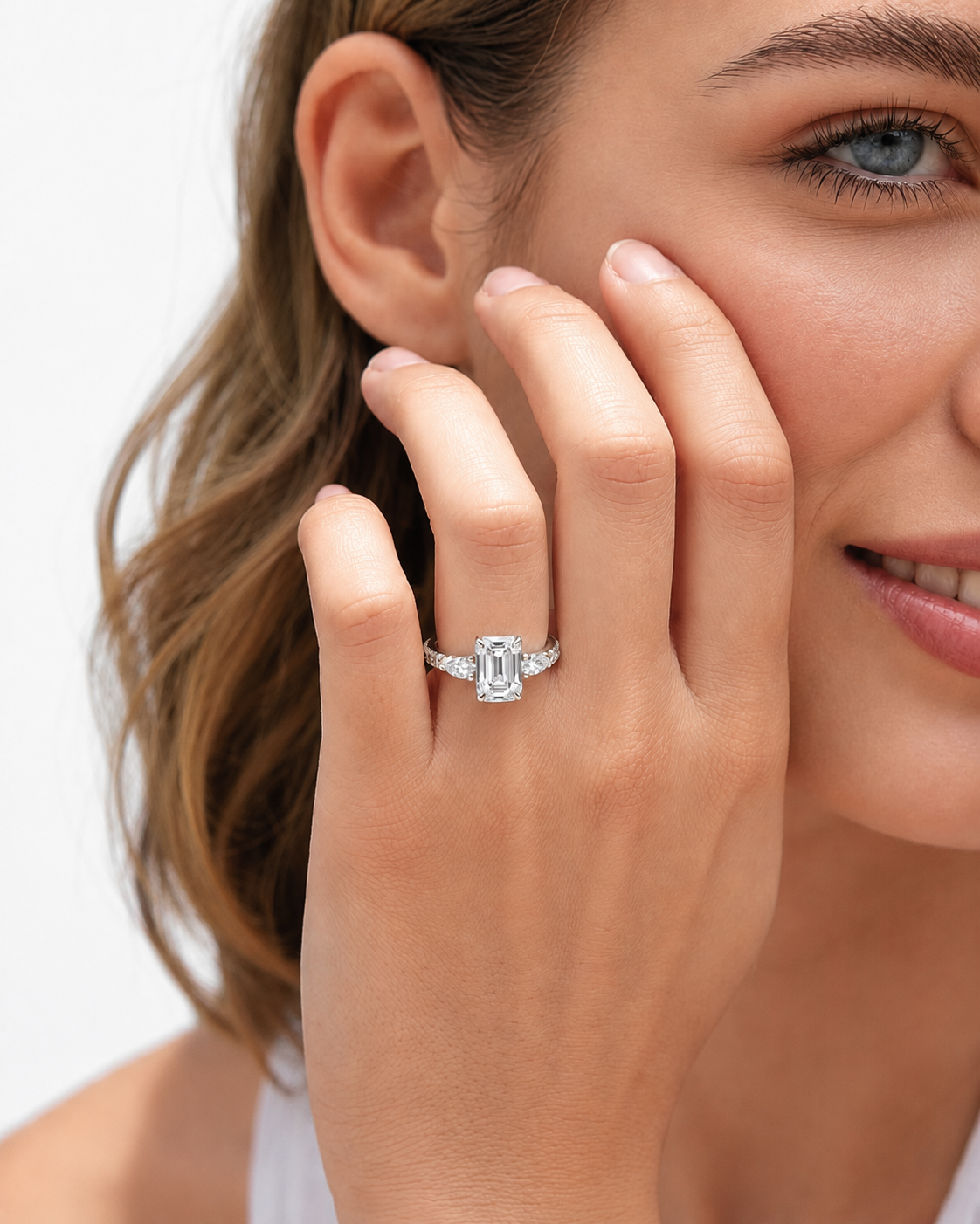 color_925_SIL,color_935_ARG,color_10K_WG, color_14K_WG &&A close-up lifestyle shot of a woman wearing a silver engagement ring with an emerald-cut center stone and smaller side stones on her ring finger.