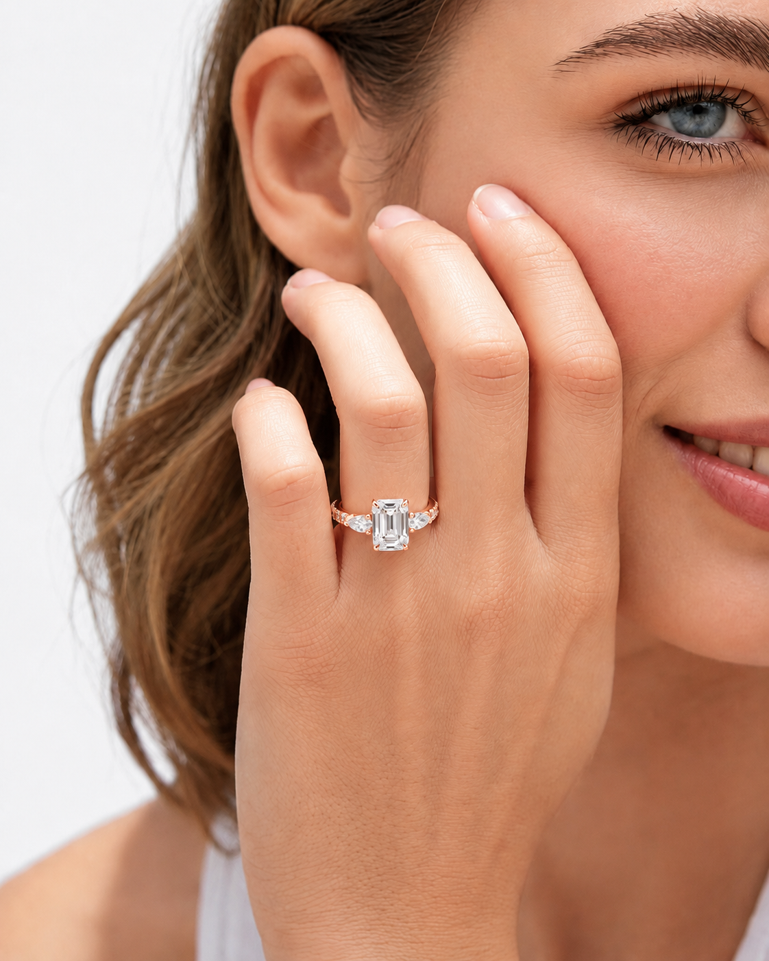 color_10K_RG, color_14K_RG &&A close-up lifestyle shot of a woman wearing a rose gold engagement ring with a rectangular emerald-cut center stone and smaller side stones, shown on her finger.