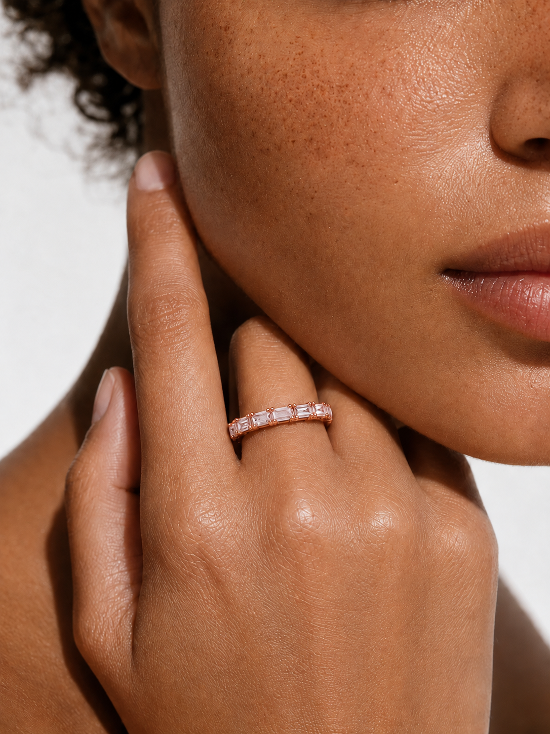 color_10K_RG, color_14K_RG &&	A close-up of a model's hand and lower face wearing a slim gold eternity ring set with small clear stones on her ring finger, against a clean white background.