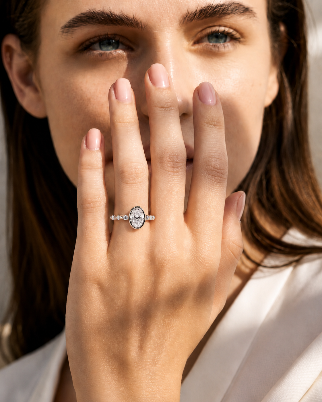 color_925_SIL,color_935_ARG,color_10K_WG, color_14K_WG &&A close-up lifestyle shot of a woman wearing a silver oval halo ring with a clear central gemstone, with her hand covering part of her face.