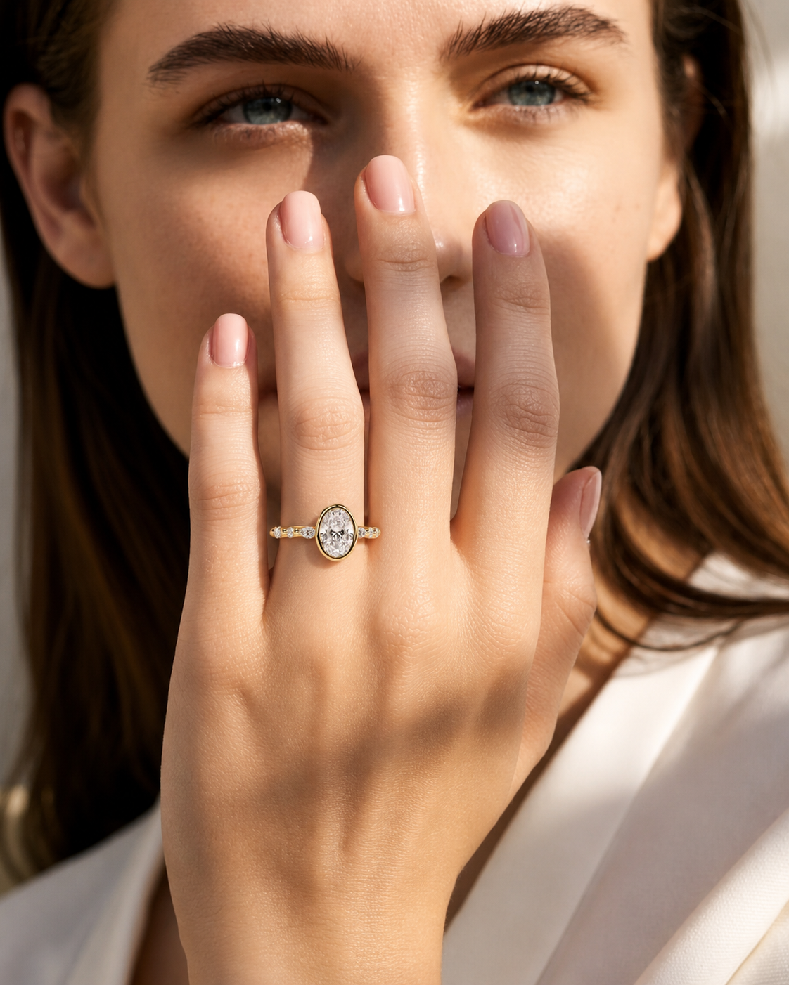 color_10K_YG, color_14K_YG &&A close-up lifestyle shot of a woman wearing a gold ring with an oval-cut clear center stone and small side stones, with her hand covering part of her face.