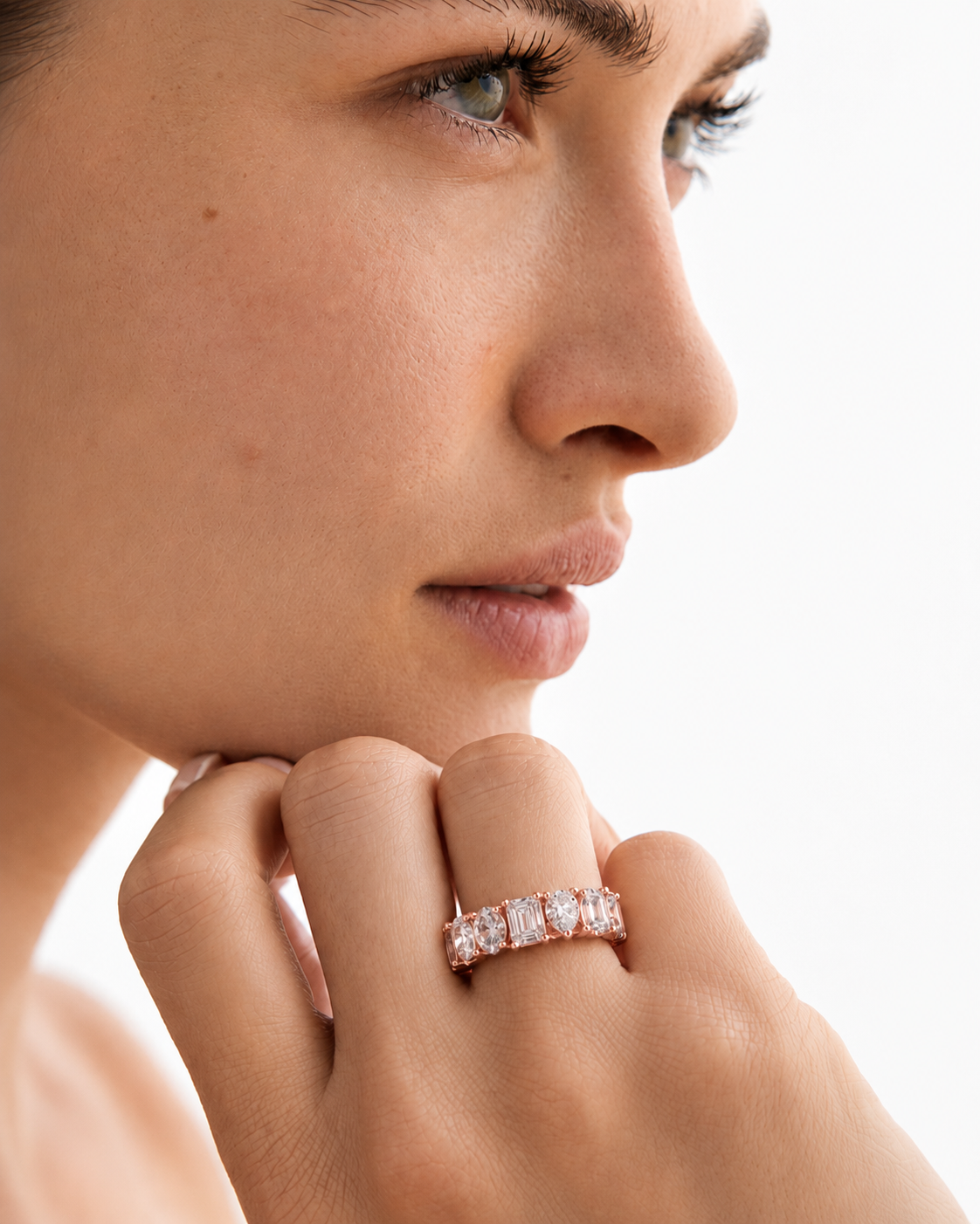 color_10K_RG, color_14K_RG &&	A close-up of a woman's face and hand wearing a rose gold eternity ring with rectangular clear stones, set against a clean white background.