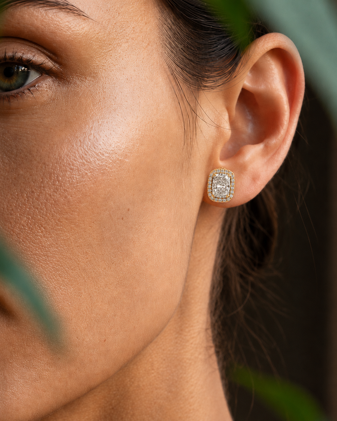 color_10K_YG, color_14K_YG &&A close-up of a woman's ear wearing a small square stud earring with a gold-toned border and clear stones, set against a softly blurred background with green leaves.