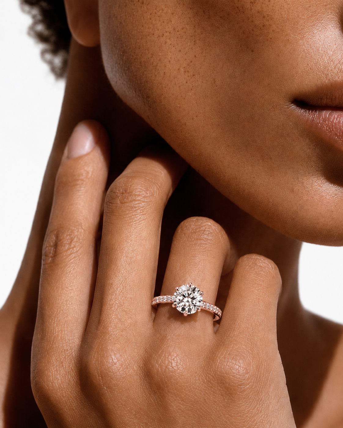 color_10K_RG, color_14K_RG &&A close-up lifestyle shot of a model wearing a diamond engagement ring on her finger, with her hand resting near her face against a clean white background.