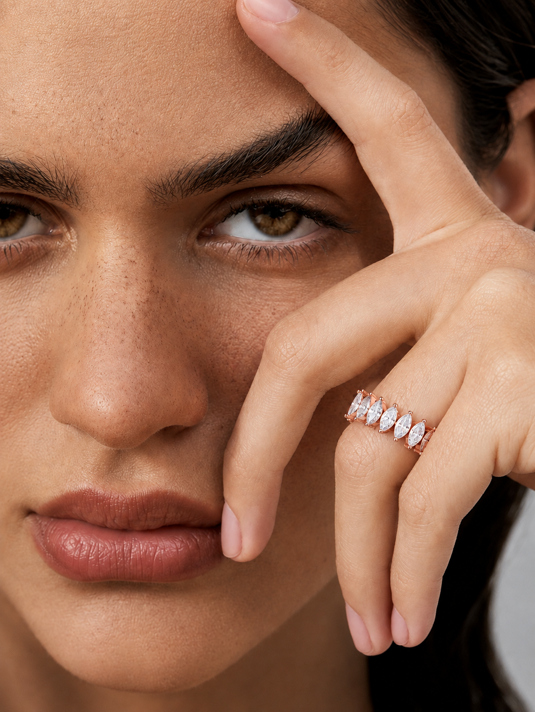color_10K_RG, color_14K_RG &&	Close-up of a woman's face and hand wearing a delicate ring with marquise-cut clear stones on a rose gold band.
