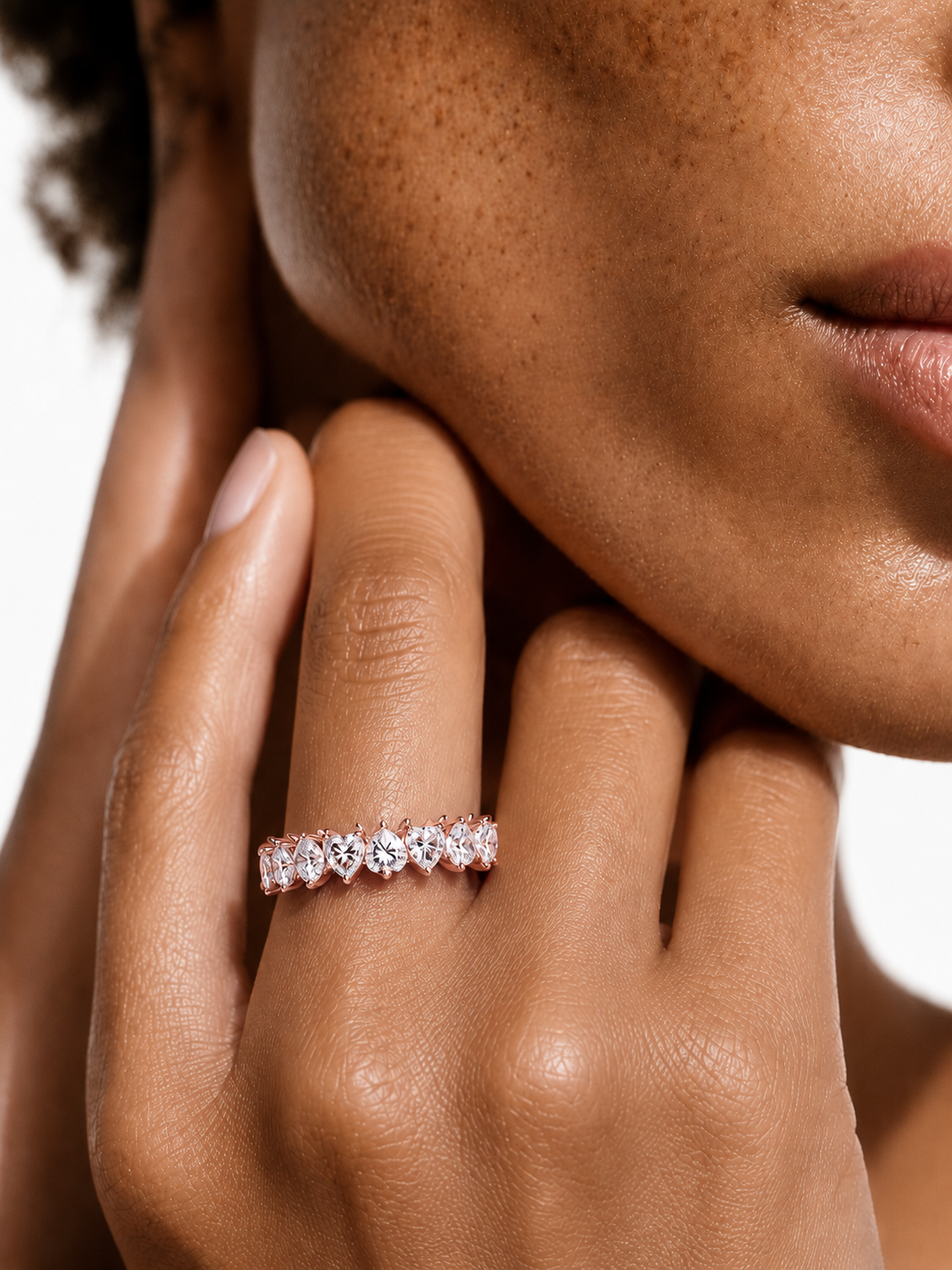 color_10K_RG, color_14K_RG &&	A close-up of a model's hand resting against her cheek, wearing a delicate rose gold eternity ring set with small clear gemstones.