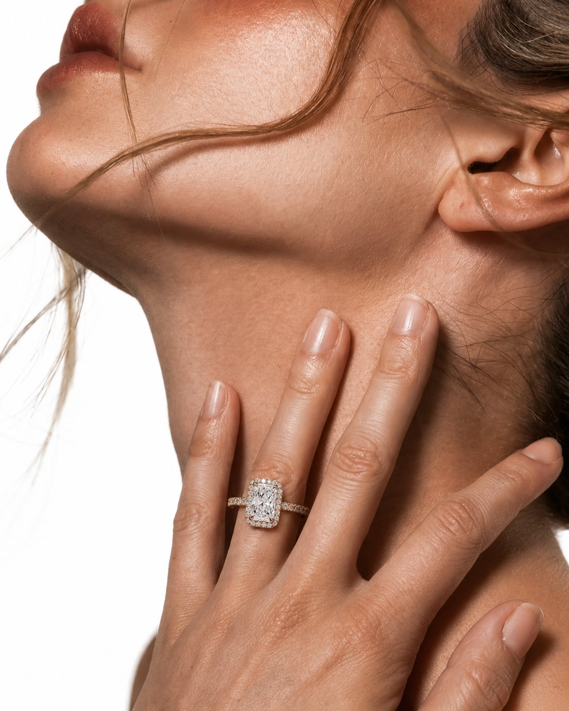 color_10K_YG, color_14K_YG &&A close-up lifestyle shot of a woman's hand resting on her neck, wearing a diamond engagement ring with a cushion-cut center stone and a pave band in a silver-toned setting.