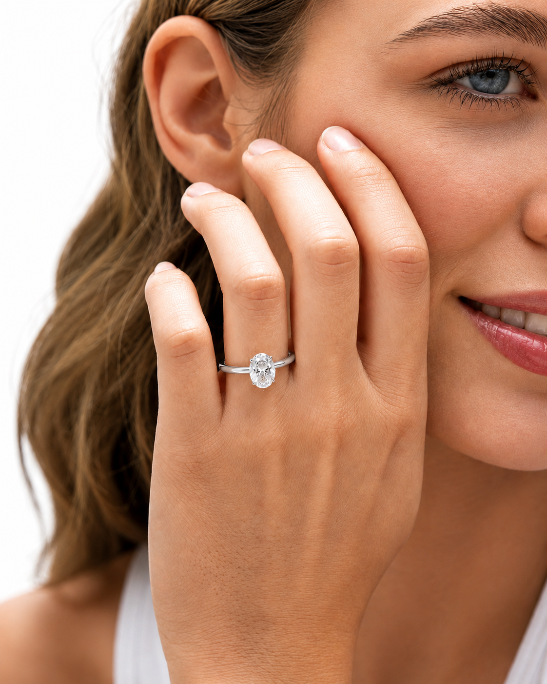 color_925_SIL,color_935_ARG,color_10K_WG, color_14K_WG &&A close-up lifestyle shot of a woman wearing a silver oval diamond engagement ring on her finger, with her hand resting against her cheek against a clean white background.