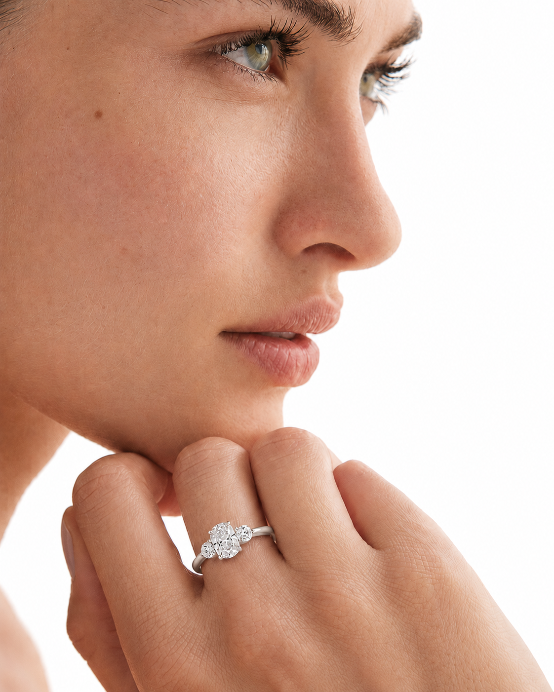 color_925_SIL,color_935_ARG,color_10K_WG, color_14K_WG &&A close-up of a woman's face and hand wearing a silver ring with a diamond cluster on her ring finger, set against a bright white background.