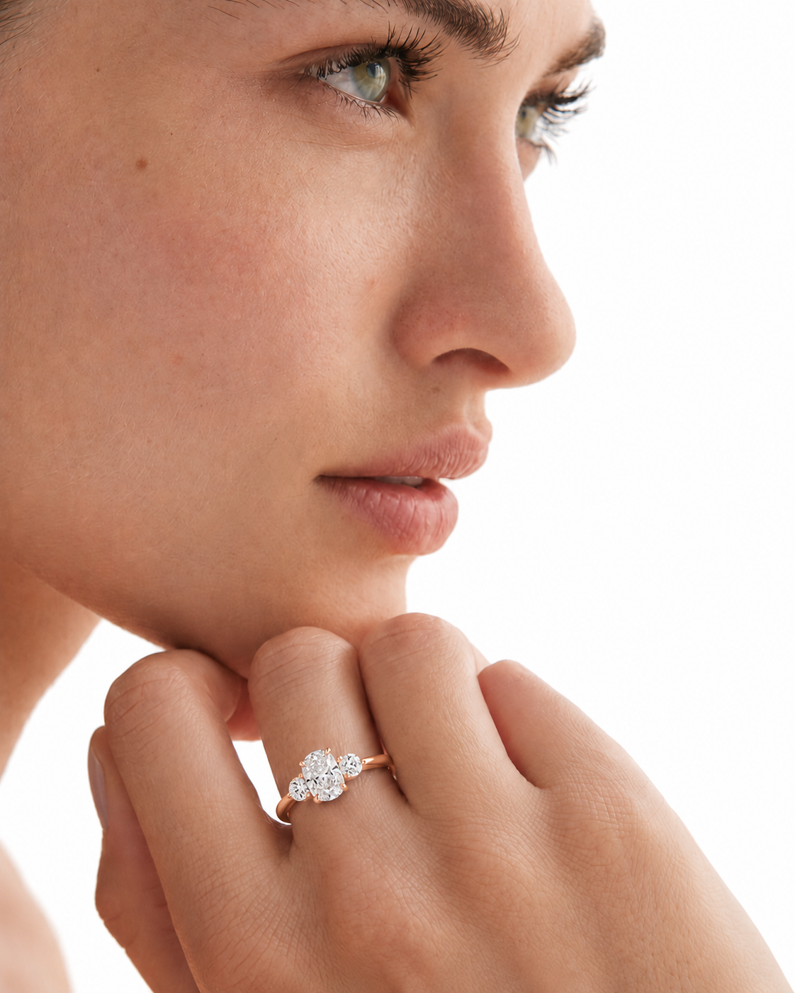 color_10K_RG, color_14K_RG &&A close-up of a woman's face and hand wearing a delicate gold ring with a small cluster of clear stones on her ring finger, set against a bright white background.