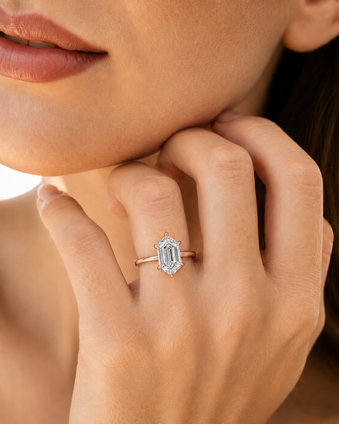 color_10K_RG, color_14K_RG &&A close-up lifestyle shot of a model wearing a rose gold ring with an emerald-cut clear center stone and a halo of small round stones.