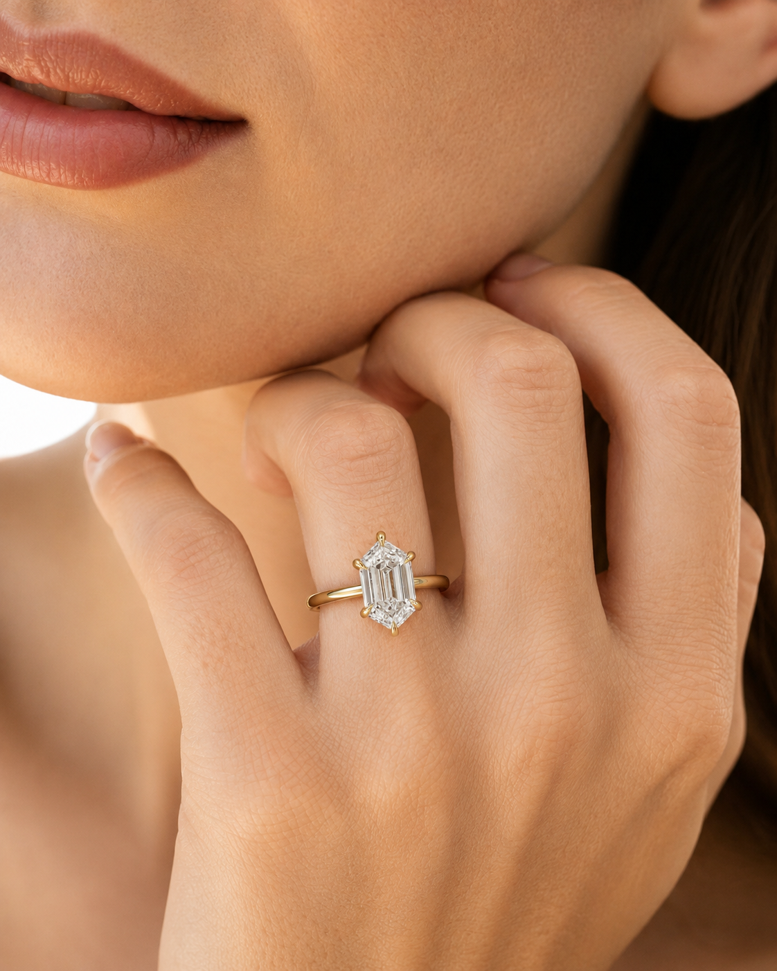 color_10K_YG, color_14K_YG &&A close-up lifestyle shot of a model wearing a yellow gold ring with an emerald-cut diamond and a small halo of round diamonds.