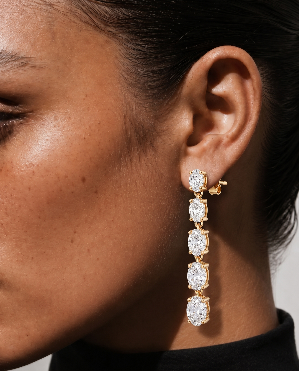 color_10K_YG, color_14K_YG &&A close-up of a model's ear wearing a gold drop earring with four round-cut clear gemstones in a vertical cascade design.