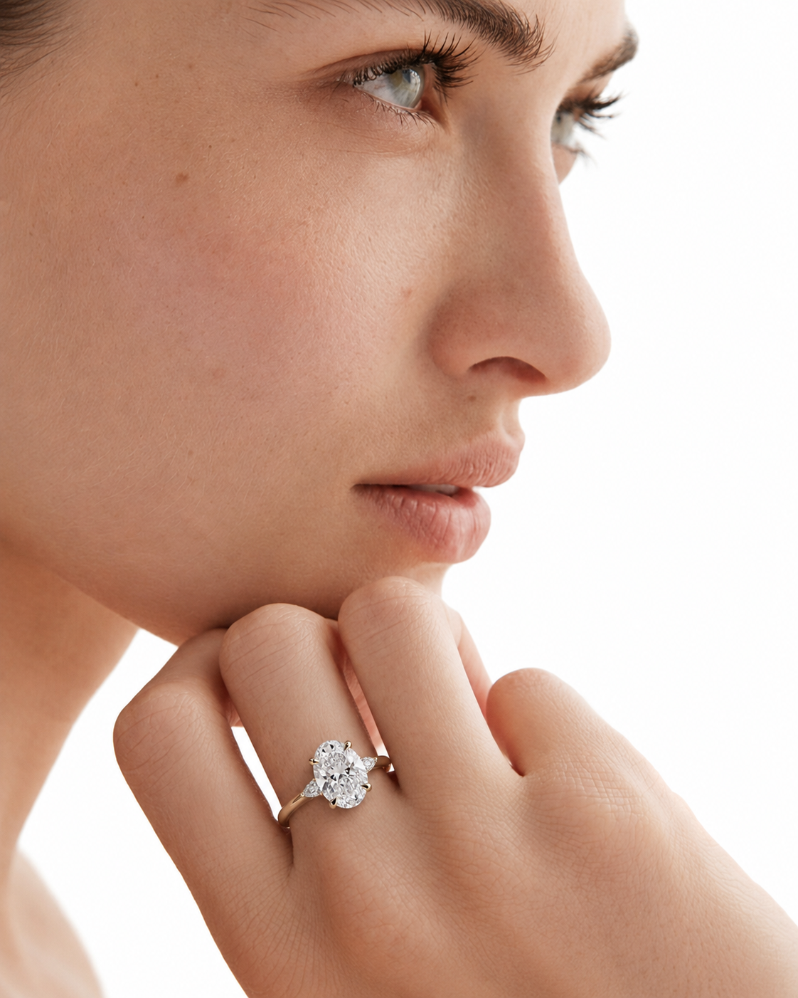 color_10K_YG, color_14K_YG &&A close-up profile of a woman resting her chin on her hand, wearing a silver-toned ring with a round-cut clear gemstone on her ring finger against a bright white background.