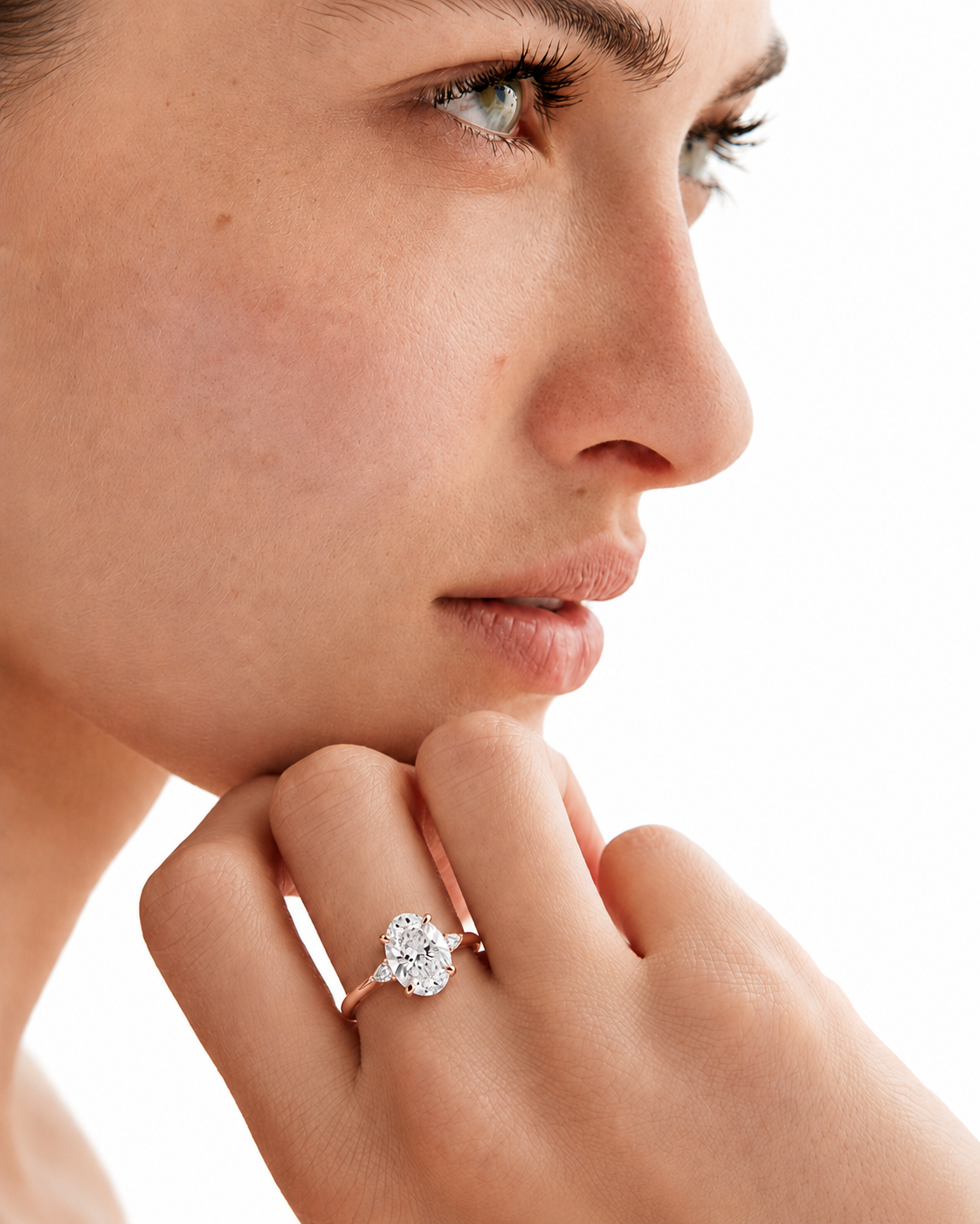 color_10K_RG, color_14K_RG &&A close-up lifestyle shot of a woman wearing a diamond engagement ring on her finger, with her hand resting near her chin against a clean white background.