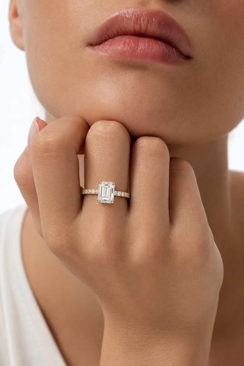 color_10K_YG, color_14K_YG &&A close-up lifestyle shot of a model wearing a silver-toned diamond ring with an emerald-cut center stone and a pave band, resting her chin on her hand against a clean white background.