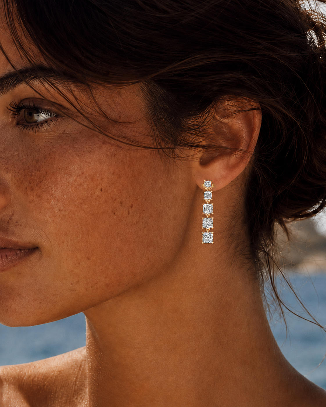 color_10K_YG, color_14K_YG &&A close-up side profile of a woman wearing a long vertical drop earring with four clear rectangular stones set in a silver-toned metal, against a softly blurred outdoor background.