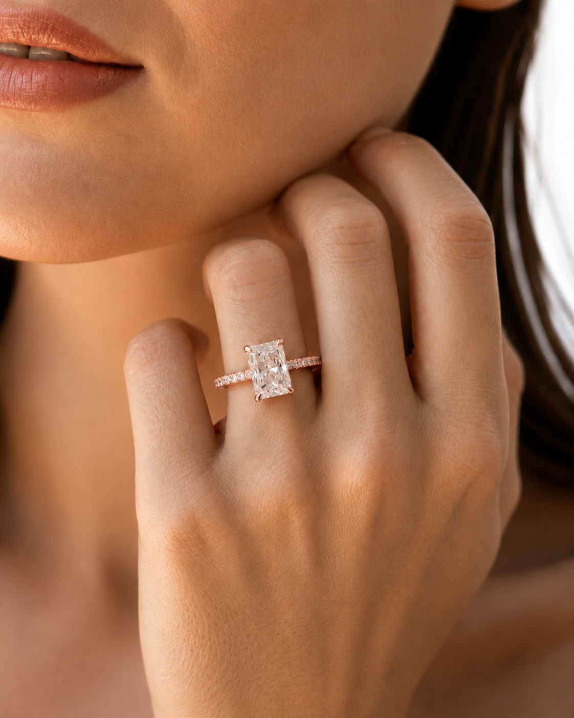 color_10K_RG, color_14K_RG &&A close-up lifestyle shot of a model wearing a rose gold engagement ring with a rectangular emerald-cut diamond. The ring is shown on the model's finger, with her hand resting near her chin against a soft, warm-toned background.