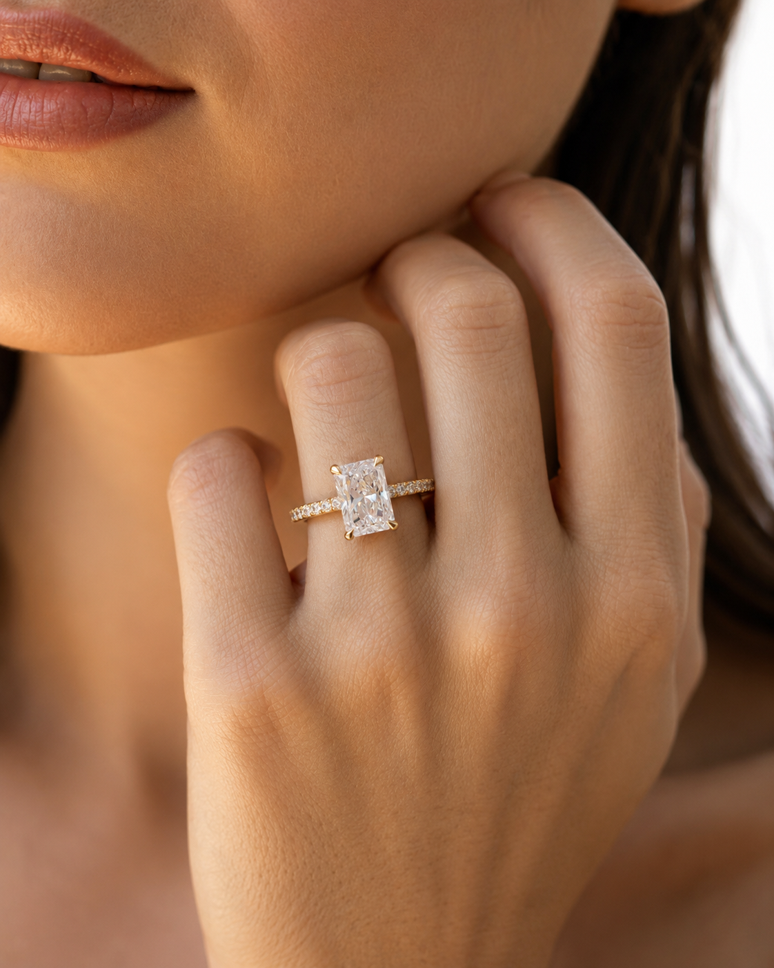 color_10K_YG, color_14K_YG &&A close-up lifestyle shot of a model's hand wearing a yellow gold ring with a rectangular emerald-cut diamond and small side stones.