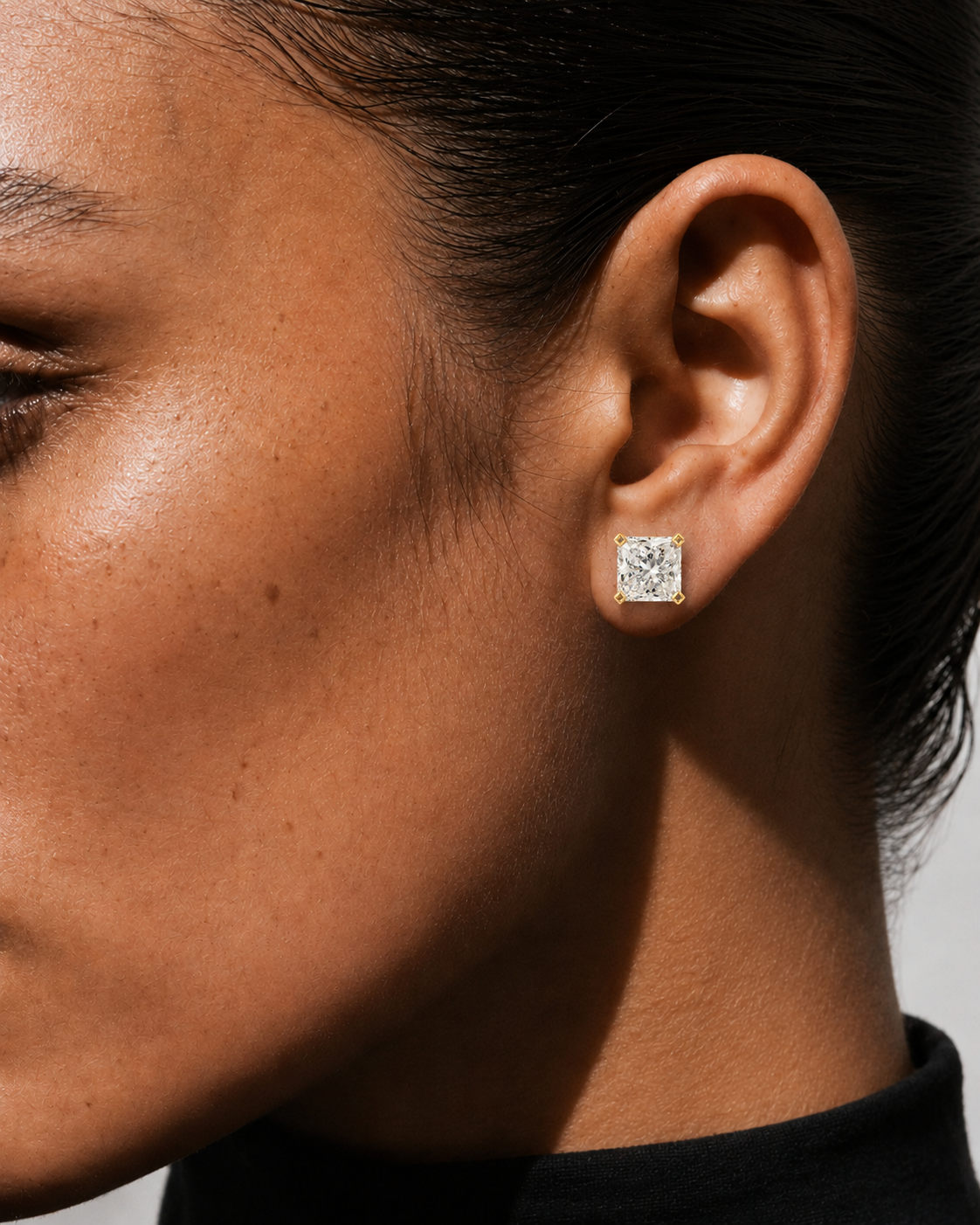 color_10K_YG, color_14K_YG &&A close-up side profile of a woman wearing a small square stud earring with a clear gemstone in a silver-toned setting.