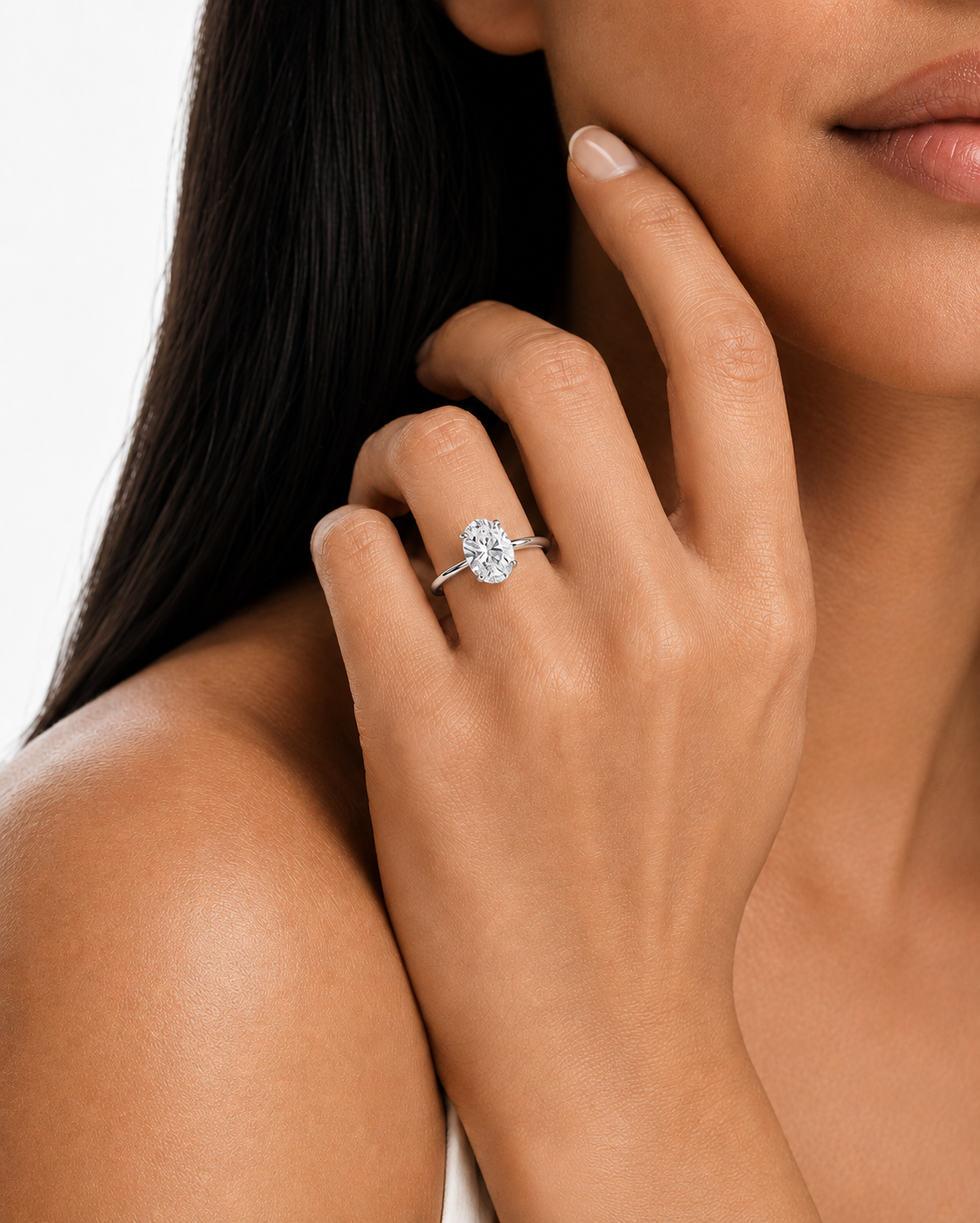 color_925_SIL,color_935_ARG,color_10K_WG, color_14K_WG &&A close-up lifestyle shot of a woman wearing a classic oval-cut diamond engagement ring on a silver band, with her hand resting near her face against a clean white background.