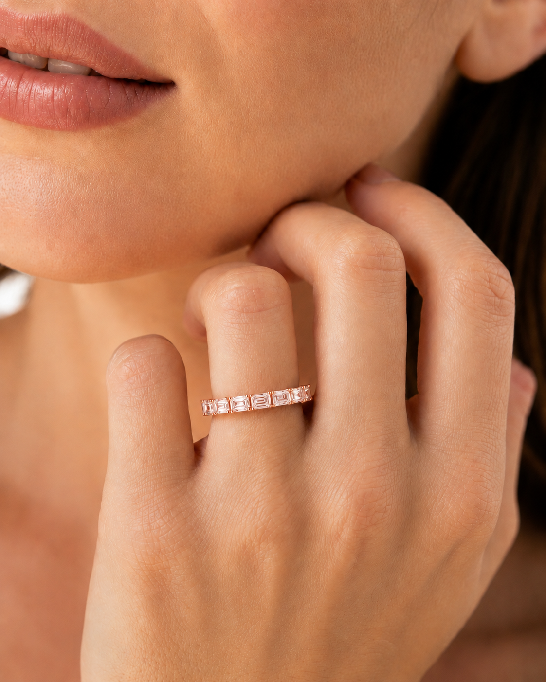 color_10K_RG, color_14K_RG &&A close-up lifestyle shot of a woman wearing a delicate rose gold eternity ring with rectangular clear stones on her ring finger.