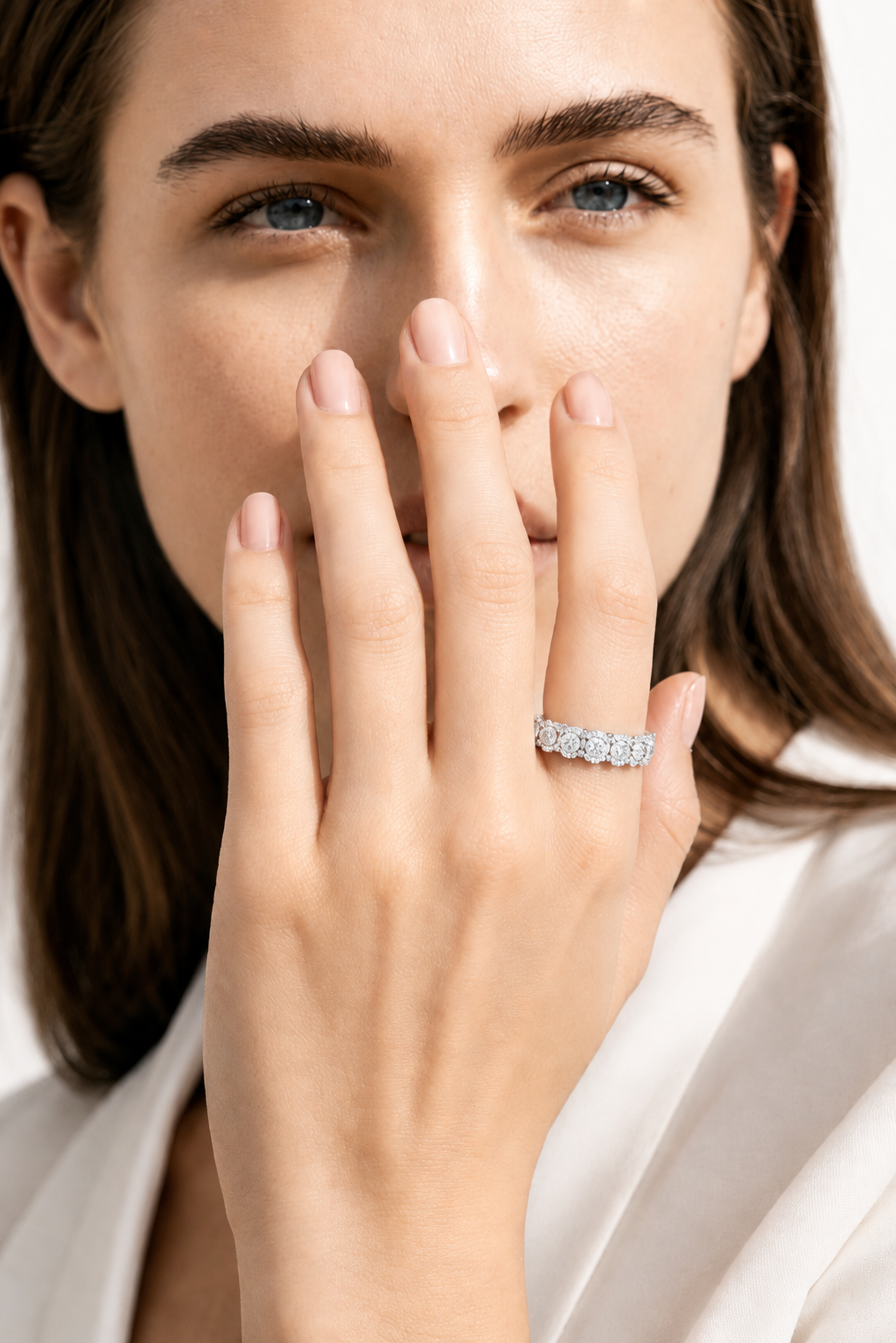 color_925_SIL,color_935_ARG,color_10K_WG, color_14K_WG &&A close-up lifestyle shot of a woman wearing a diamond eternity ring on her finger, with her hand covering part of her face. She is wearing a white satin blouse against a clean white background.