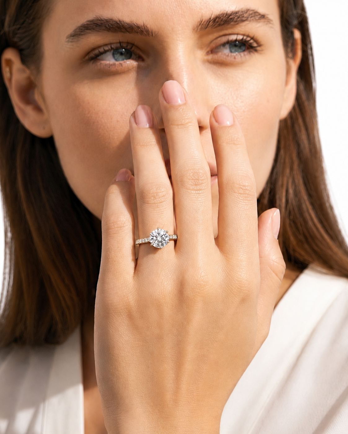 color_925_SIL,color_935_ARG,color_10K_WG, color_14K_WG &&A close-up lifestyle shot of a woman wearing a diamond engagement ring on her finger, with her hand covering part of her face against a bright white background.