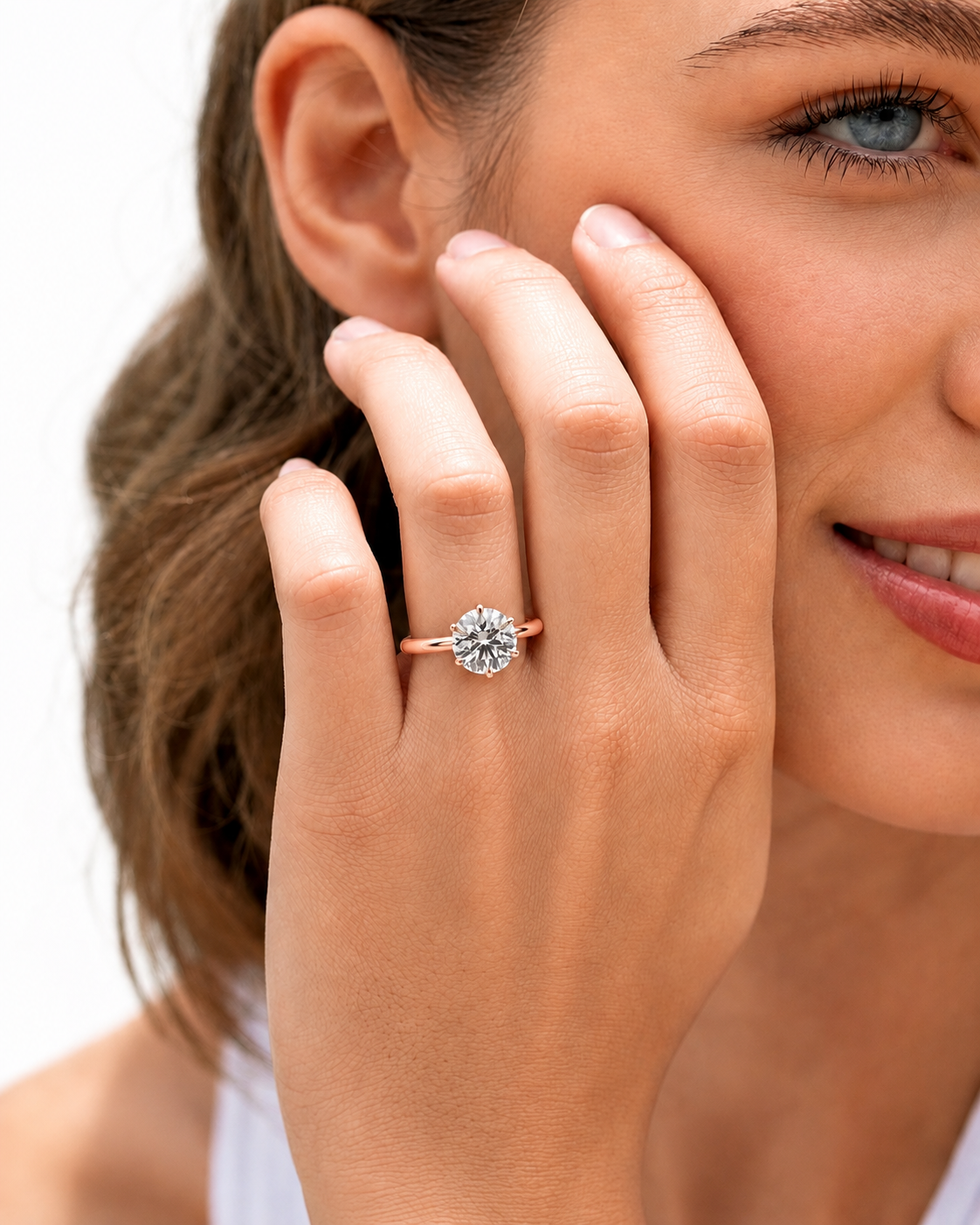 color_10K_RG, color_14K_RG &&A close-up lifestyle shot of a woman wearing a rose gold engagement ring with a round-cut clear gemstone on her finger, with her hand resting against her cheek.