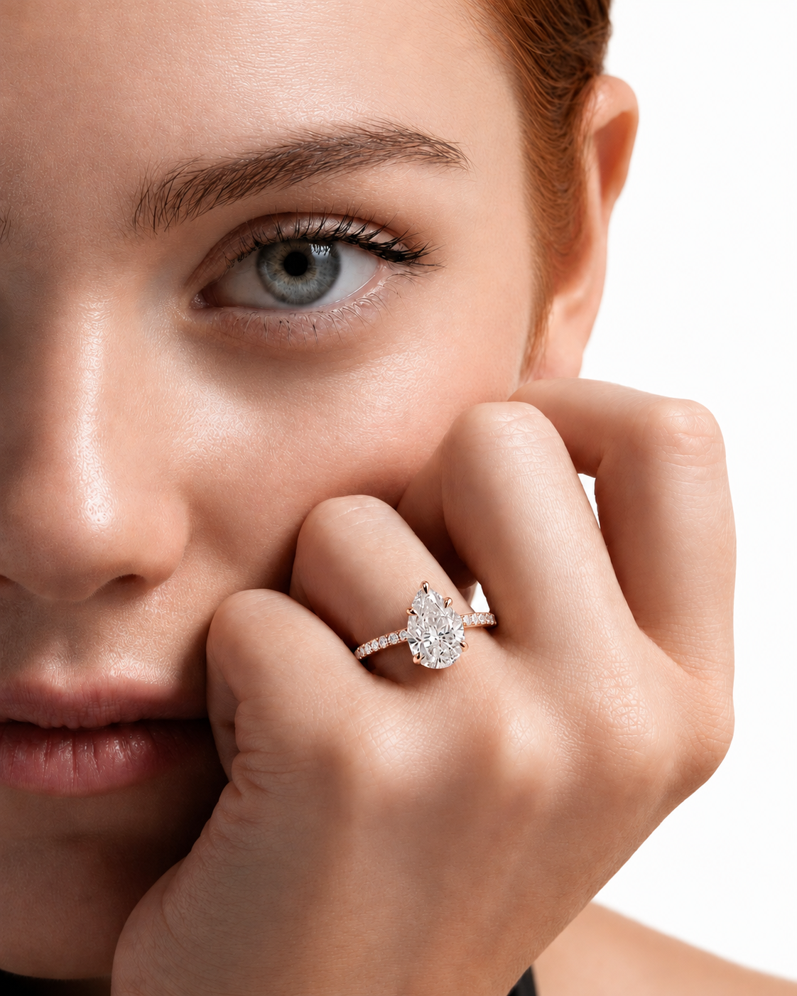 color_10K_RG, color_14K_RG &&A close-up lifestyle shot of a model wearing a pear-cut diamond engagement ring on her finger, with her hand resting near her face against a clean white background.