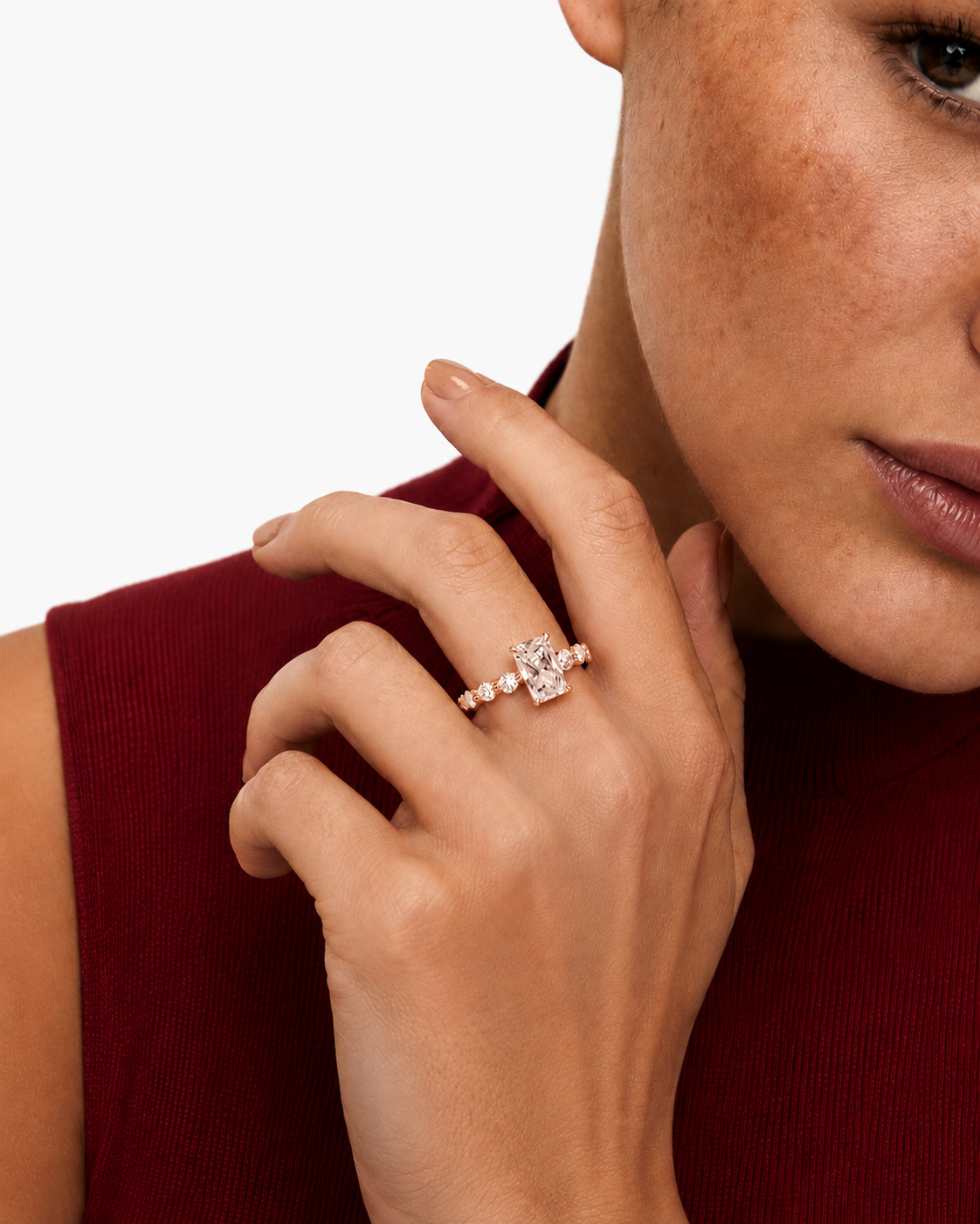 color_10K_RG, color_14K_RG &&A close-up lifestyle shot of a model wearing a rose gold ring with a central clear gemstone and small side stones on her finger. She is wearing a dark red ribbed top against a clean white background.
