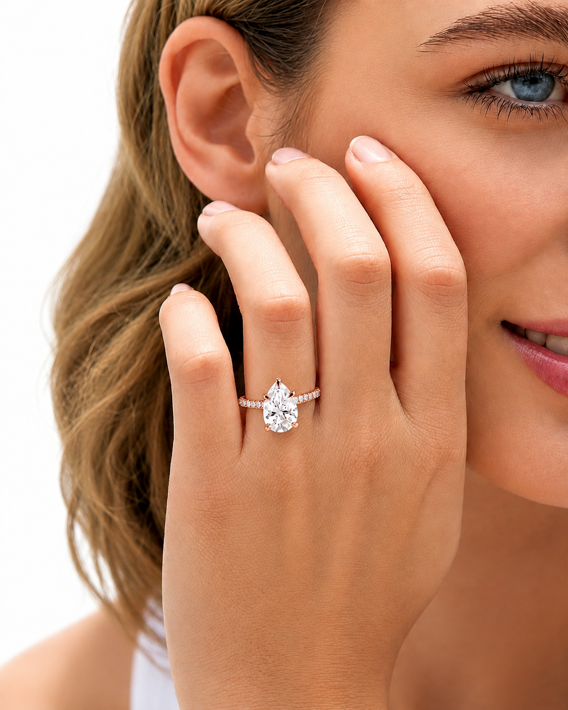 A close-up lifestyle shot of a woman wearing a pear-cut diamond engagement ring on her finger, with her hand resting against her cheek against a clean white background.
