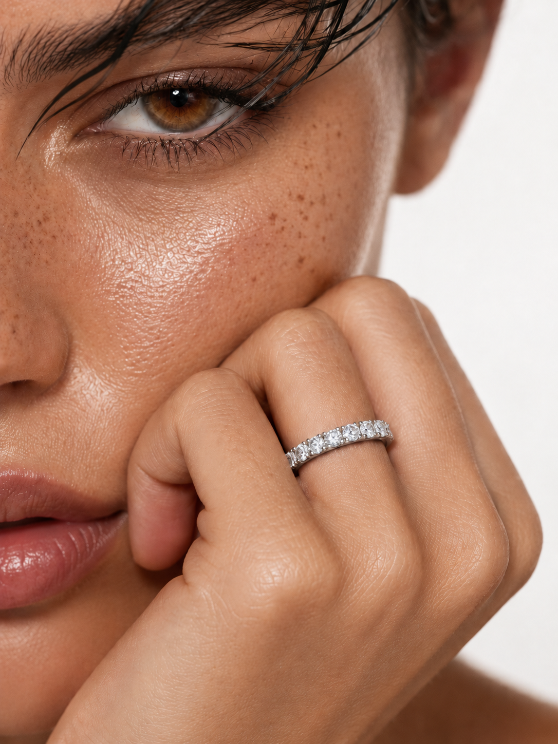 color_925_SIL,color_935_ARG,color_10K_WG, color_14K_WG &&A close-up lifestyle shot of a model wearing a sparkling diamond eternity ring on her finger, with her hand resting near her face against a clean white background.