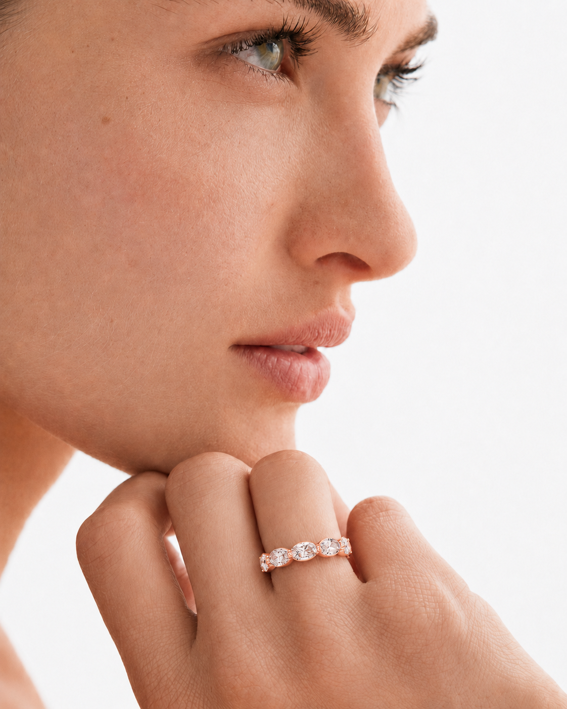 color_10K_RG, color_14K_RG &&A close-up lifestyle shot of a woman's face and hand, wearing a delicate rose gold band ring set with small round clear gemstones.