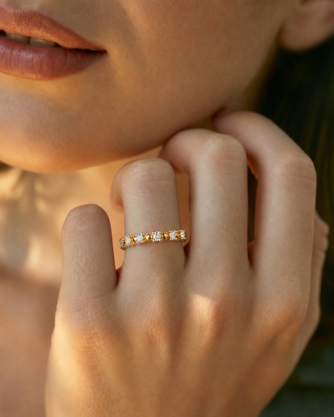 color_10K_YG, color_14K_YG &&A close-up lifestyle shot of a model's hand wearing a delicate gold eternity ring set with small round-cut clear gemstones, with warm sunlight casting soft shadows.